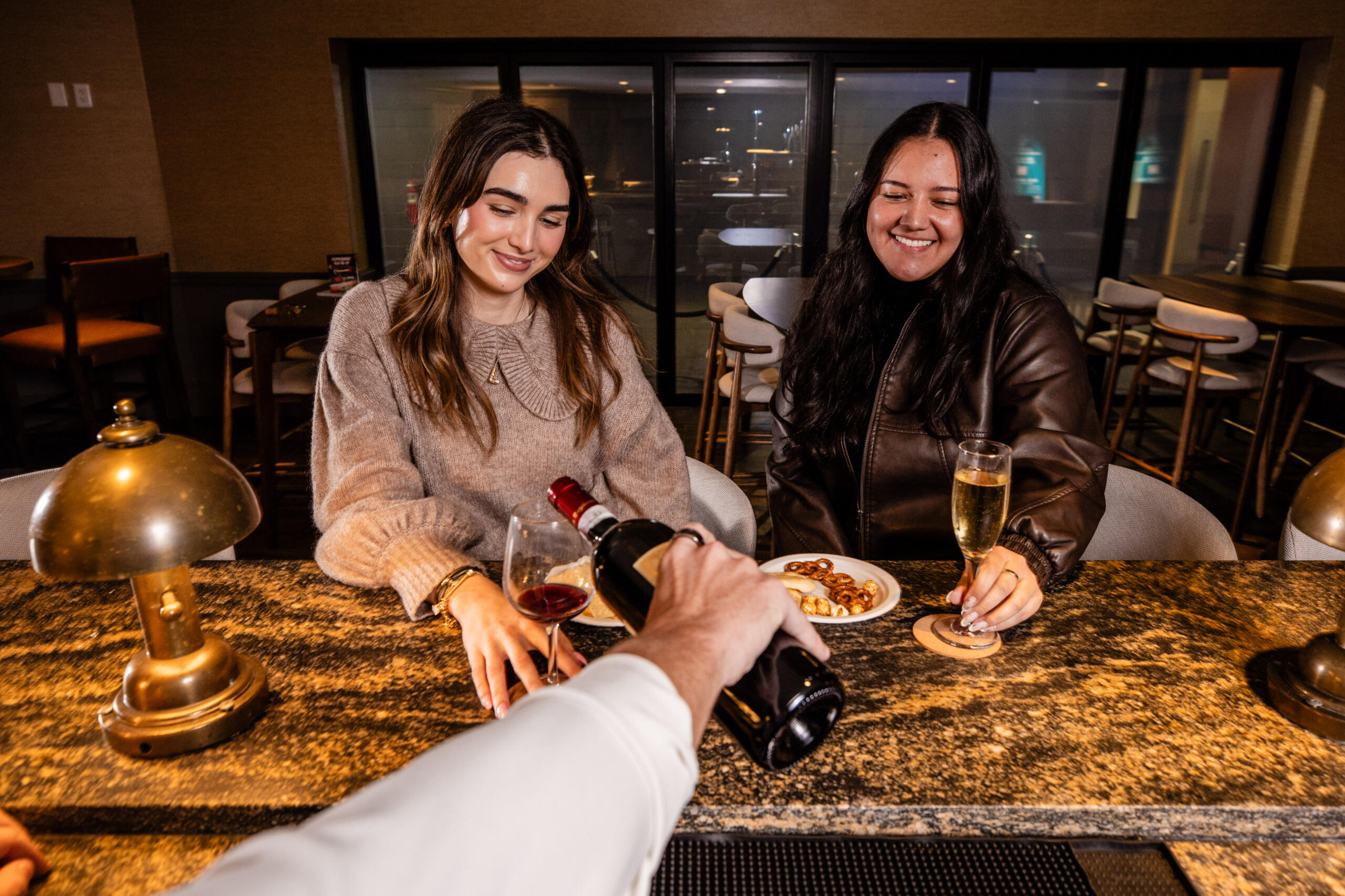 Image of two friends smiling by the bar having their wine refilled by the bartender