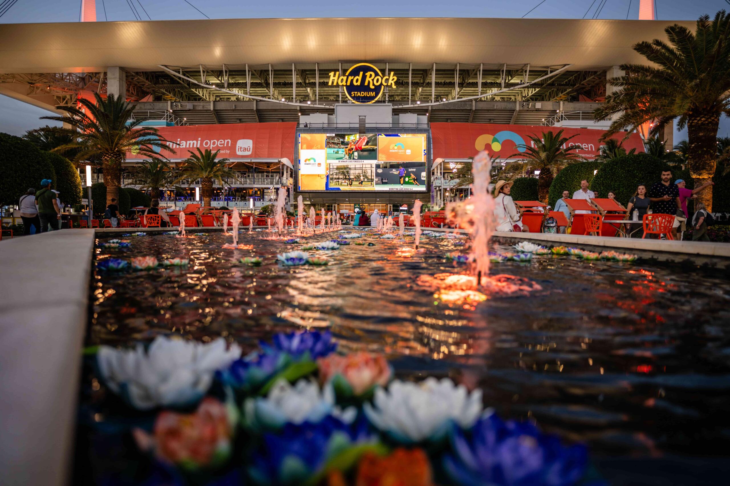 image of the fountain at night with colorful flowers floating. Stadium lights are seen in the background.