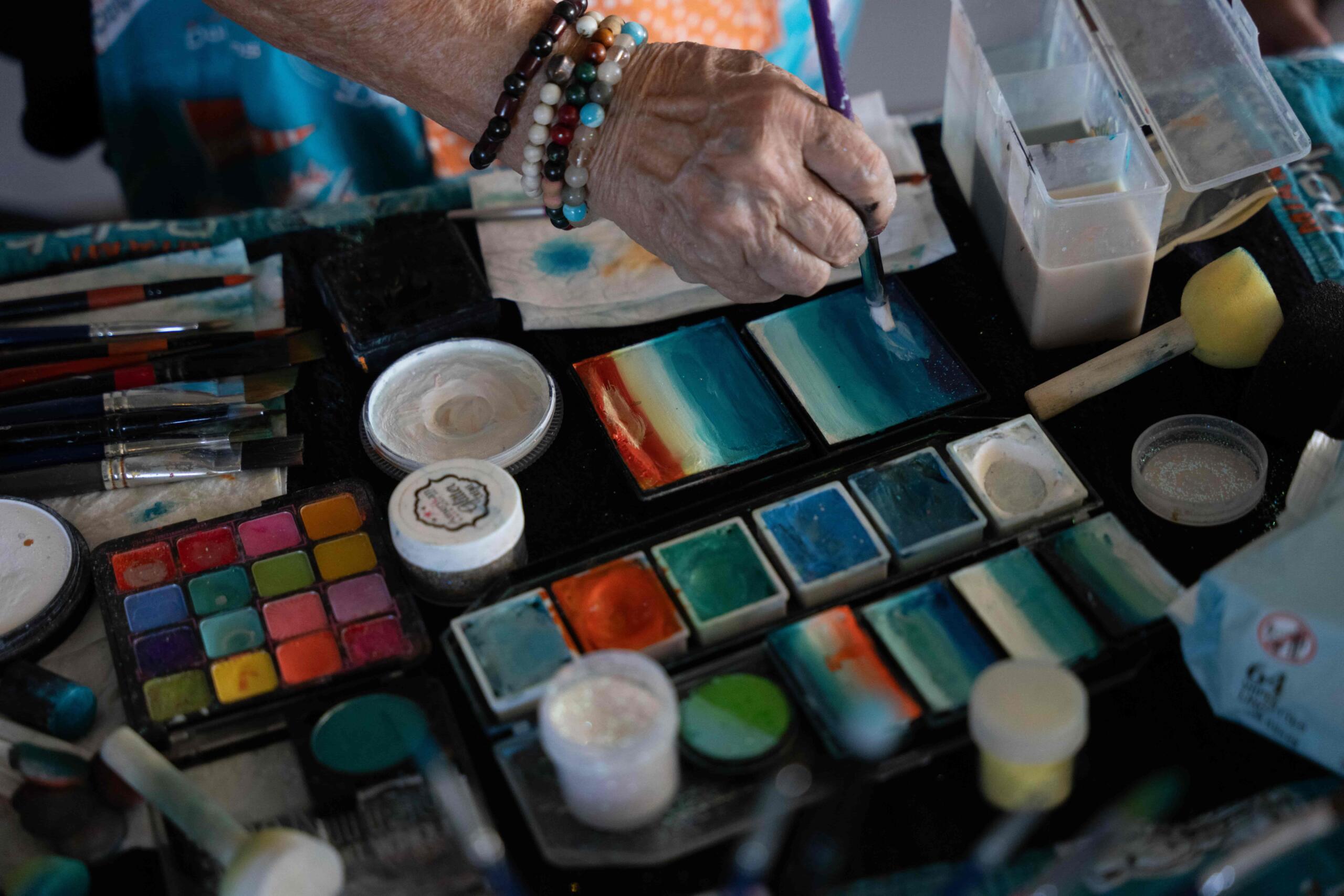 Artist hand holds paintbrush and all their art material is layed on the table. There are many colors.